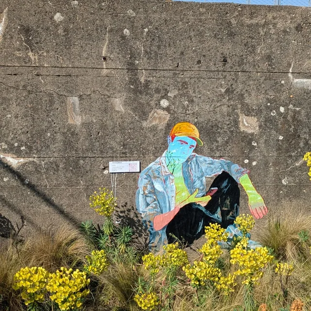 Collage sur un mur de béton, personnage penché au-dessus d'un massif de fleurs jaunes.