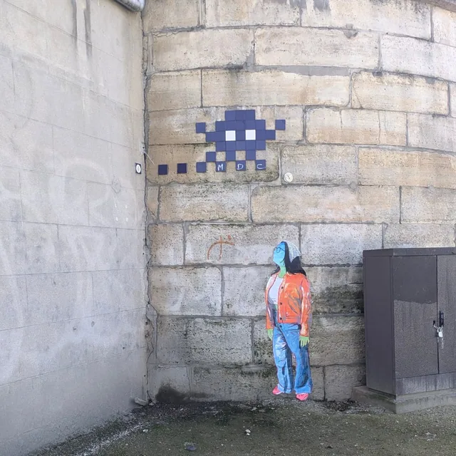 Collage sur un mur de pierre, petit personnage debout sous une mosaïque en forme d'extraterrestre.