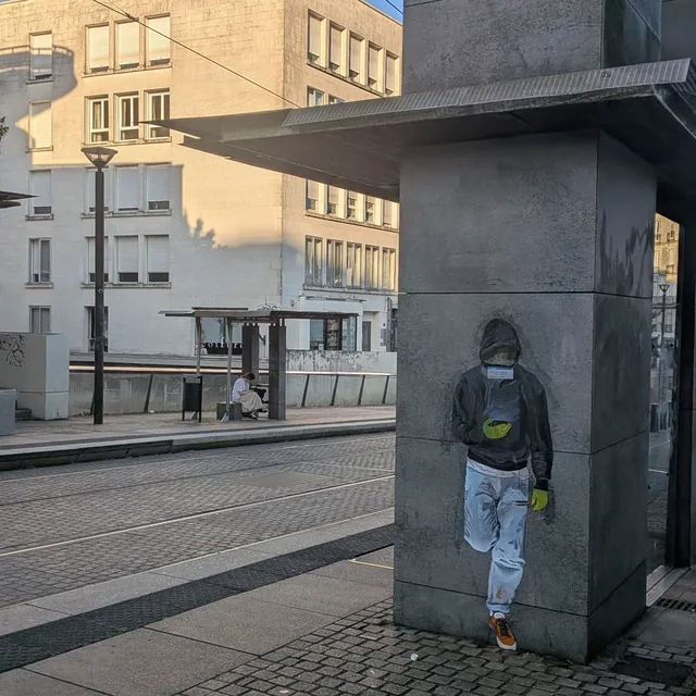 Collage sur un pilier de béton en ville, personnage debout la capuche relevée.