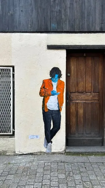 Collage sur la façade d'une maison, personnage adossé au mur près d'une porte en bois.