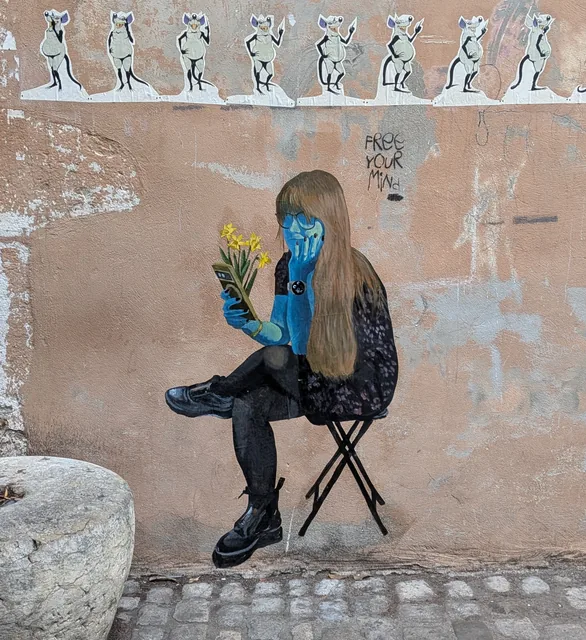 Collage dans la rue : une jeune femme à la peau bleue, assise sur un tabouret pliant, regarde son téléphone en tenant des jonquilles, devant un mur orangé écaillé.