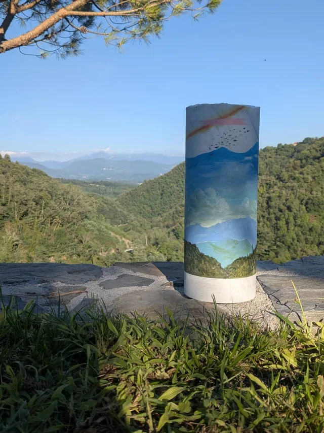 Cylindre de marbre peint d'un paysage de mer et de ciel, posé sur un socle face à des collines boisées.